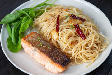 Spaghetti olio with salmon steak and herbs in white plate on dark wooden table background.