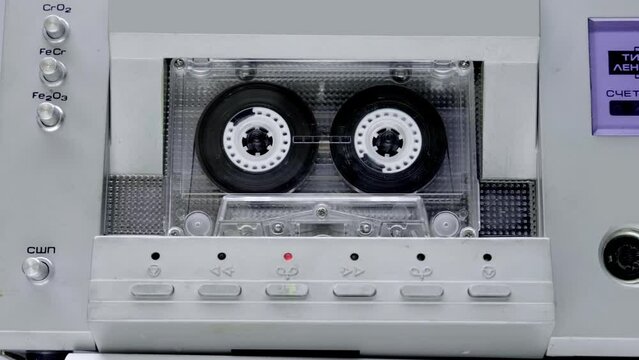 A cassette with transparent body plays music in a Hi-Fi deck from 80s of silver  color. Closeup. Shot in motion