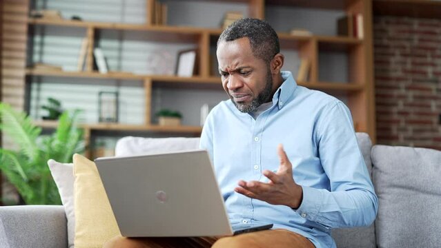 Frustrated mature african american man having problems with software or service on laptop computer sitting on sofa in room at home. Worried disappointed male complains about a bad internet connection