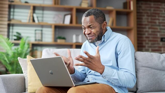 Angry dissatisfied mature african american man arguing on a video call using laptop sitting on sofa in living room at home office. Excited businessman emotionally talks with subordinate about problems