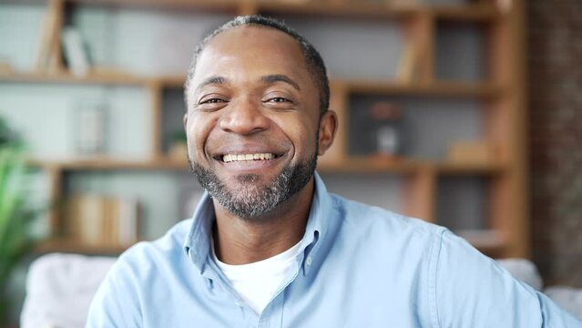 Portrait of happy mature African American male in shirt sitting on sofa in living room at home office. Head shot of a positive businessman. A smiling freelancer poses looking at the camera. Close up
