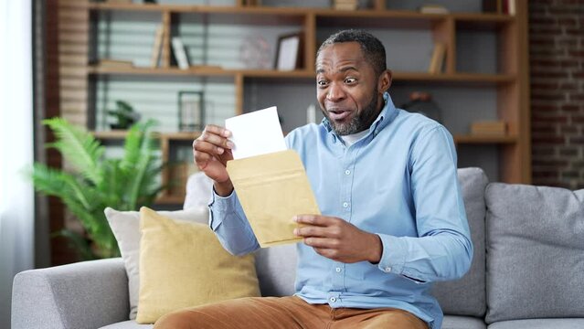 Happy excited mature african american man reading letter with great news sitting on sofa in living room at home. Joyful senior bearded male is satisfied with pleasant notification, celebrates success