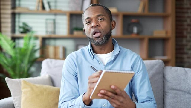 Webcam view. Mature african american male watching video call online training notes in notebook looking at camera sitting in home. Senior bearded black man listening remote learning talking with tutor
