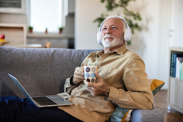 Senior man wearing headphones to listen to music on a laptop on couch at home. Retired male...