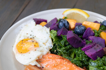 Healthy salad with fried egg, salmon, kale, sweet potato and blueberries on white plate.