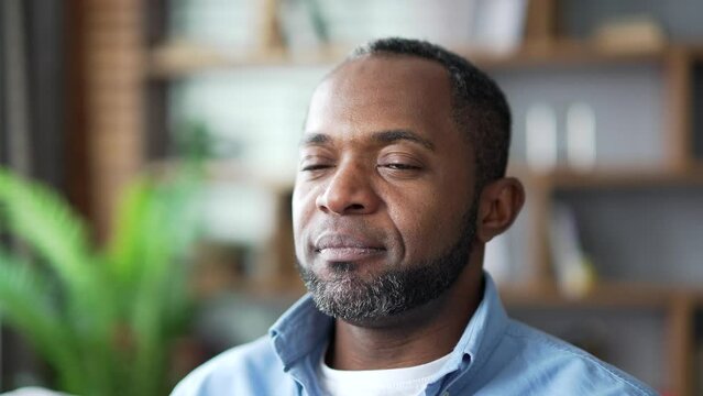 Close up of a mature african american man relaxing with his eyes closed while sitting on sofa in living room at home. Face of calm happy senior bearded male resting after work and looking at camera
