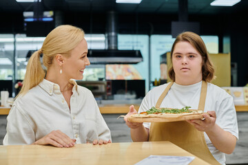 young waitress with mental disability holding delicious pizza near joyful woman sitting in cafe