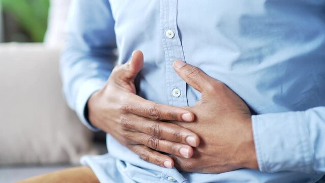 Close up. Male hands holding massaging stomach. Mature african american man in blue shirt feels stomach pain sitting on sofa in living room at home. Sick man suffers from bloating, gastritis or spasms