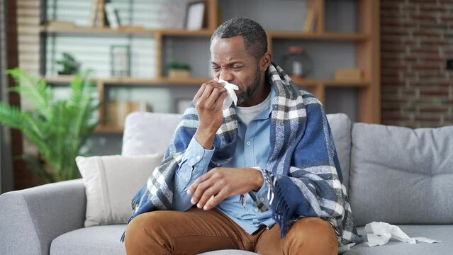 Sick mature african american male suffers from a runny nose sitting on sofa in the living room at home. Senior bearded black man wipes his nose with a handkerchief. He has a cold, fever, virus or flu