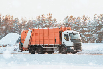 An orange sanitation truck moves on a snow-covered winter road. Garbage truck outdoor. Recycling truck rides on the road.