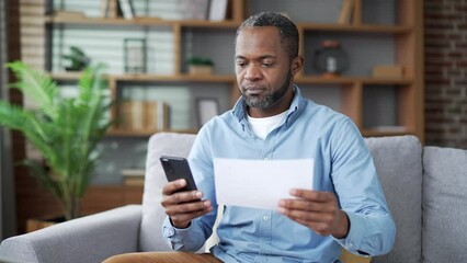 Mature african american man uses mobile app for spending bills using smartphone while sitting in room at home. Male calculates household finances and enters data into a chat bot easy and simple to pay - Powered by Adobe