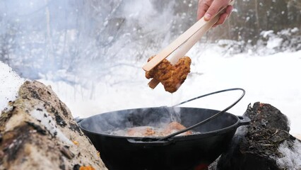 Close up of male hiker cooking food on open bonfire with hiking kitchen utensils on snowy background. Preparing hearty dishes during meal break while having outdoor activities in wild nature.