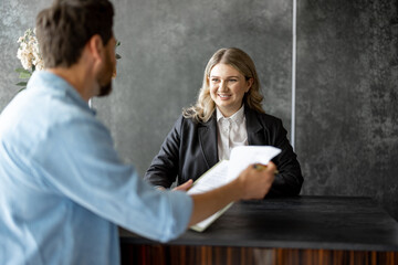 Young medical receptionist talks with a male client and gives some paper to sign at the reception desk of modern clinic - Powered by Adobe