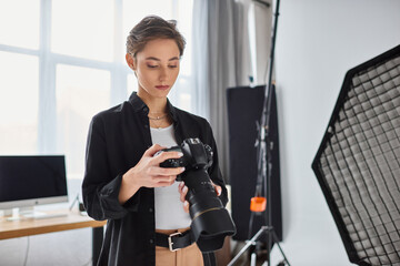 young attractive female photographer in casual outfit looking at photos on her camera in studio - obrazy, fototapety, plakaty