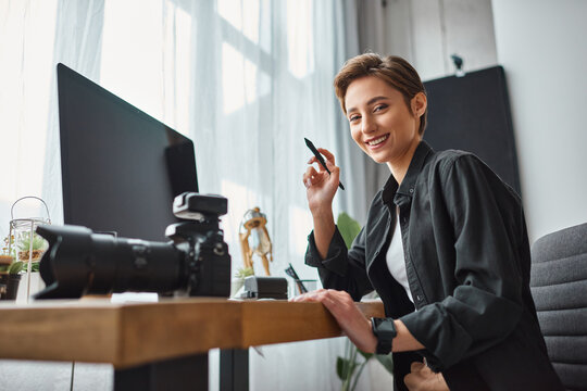 cheerful woman working with drawing tablet and her camera in front of computer and smiling happily