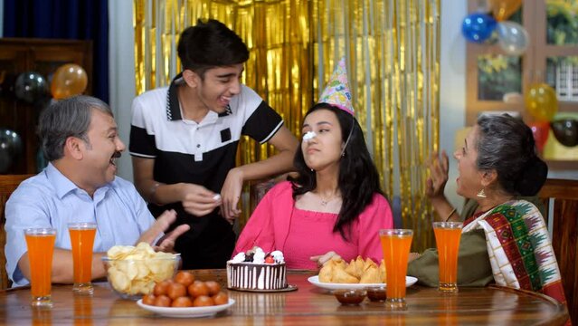 A teenage boy feeding and smudging her sister's face with a cake - angry sister  elder sister  sibling bonding  fun and enjoyment. A beautiful girl celebrating her birthday with her parents - two g...