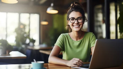 Foto op Plexiglas Cheerful professional woman wearing glasses and a green blouse is sitting at a desk with a laptop and holding coffee cup © MP Studio