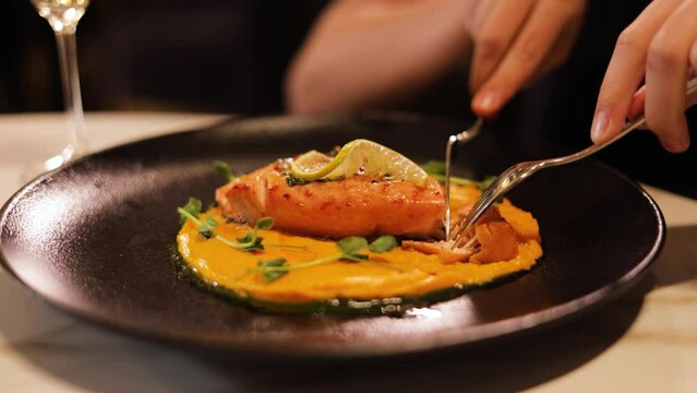 Close up of woman sitting at table at restaurant and cutting seafood dish with fork and knife in elegant way. Grilled salmon fillets served on black round plate with potato puree and pea sprouts.
