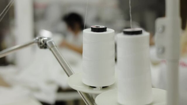 Close up of white spools of thread getting used in sewing process on blurred industrial background. Automation of textile production in factories for working condition facilitating of employees.