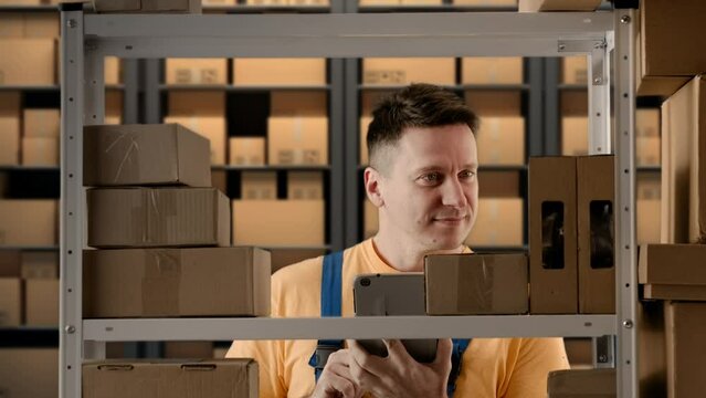 Portrait of male working in storage. Man storekeeper standing near rack with boxes, checking good writing on tablet, positive face.