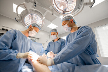 Three confident surgeons performing surgical operation on a patient's knee in operating room. Concept of real surgery and invasive treatments - obrazy, fototapety, plakaty