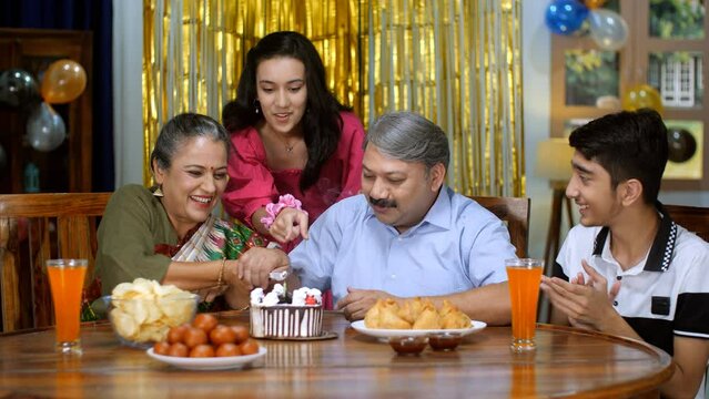 A middle-aged Indian husband and wife blowing a candle and cutting a cake together - Indian couple marriage anniversary  celebration time. An old Indian couple celebrating a birthday with their gra...