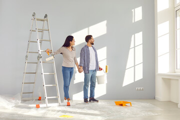 Smiling happy couple painting the wall of their new home holding paint rollers and looking at the window near ladder. Married man and woman doing repair renovation preparing to move into a new flat. - obrazy, fototapety, plakaty