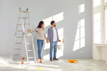 Foto op Canvas Smiling happy couple painting the wall of their new home holding paint rollers and looking at the window near ladder. Married man and woman doing repair renovation preparing to move into a new flat. © Studio Romantic