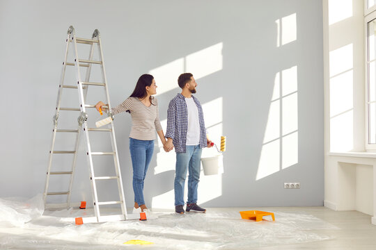 Smiling happy couple painting the wall of their new home holding paint rollers and looking at the window near ladder. Married man and woman doing repair renovation preparing to move into a new flat.
