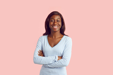 Portrait of happy woman in casual wear. Young African American girl light blue long sleeve top smiling, holding her arms folded and looking at camera isolated on pastel pink background