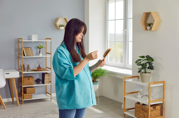 Upset young pregnant woman in casual clothes standing in living room at home and holding a comb...