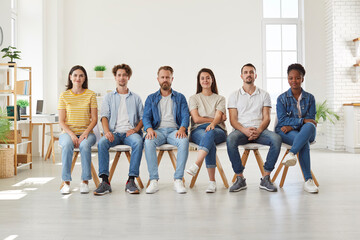 Different multiracial young people in casual clothes sit in row on chairs and look at camera. Group of positive men and women in jeans and t-shirts sitting together on chairs in room. Team concept.