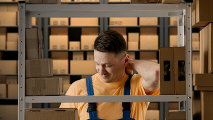 Portrait of male working in storage. Man storekeeper standing near rack with boxes tired face massaging neck has back pain after work.