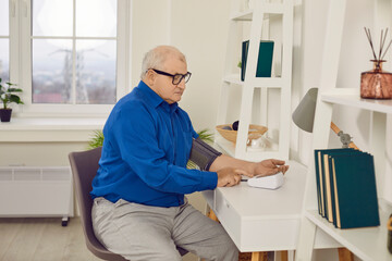 Senior man measuring arterial blood pressure himself sitting at the desk at home. Elderly retired person checking high blood pressure. Healthcare and medicine and hypertension problem concept.