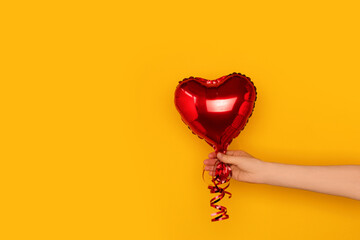 Hand hold red inflatable foil balloon in a heart shape. Festive concept in front of yellow background. - obrazy, fototapety, plakaty