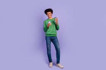 Full length photo of positive cheerful man dressed green sweater drinking tea typing modern device empty space isolated violet color background