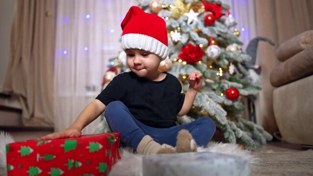 Adorable kid in Santa cap claps his hand by the gift boxes. Baby boy sits on the floor near the Christmas tree.