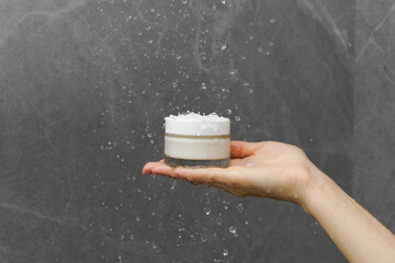 Female hand holding white mockup jar of face and body cream or oil under water drops in shower on...