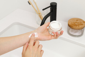 Female fragile hands apply white cream from a glass jar on the wrist on the background of the sink...