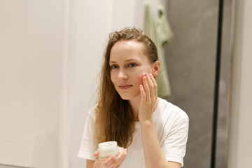 A young woman applies moisturizer to her facial skin while looking into the mirror in the bathroom. Beauty care concept