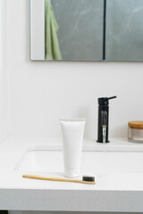 A white mockup of a tube of toothpaste and a wooden toothbrush stand on the sink the background of...
