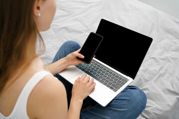 Naklejka premium Young woman holding phone and laptop with blank black screen while sitting on sofa. Background mockup for your advertising content. The concept of remote work and watching online courses and movies.
