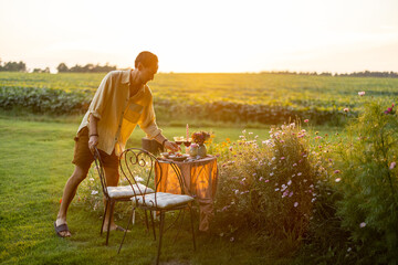 Man serving table for romantic dinner on a green lawn with flowers during a sunset
