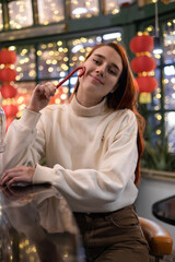 Beautiful girl dressed in trendy clothes with candy cane sitting in a cafe decorated for Christmas and Chinese New Year. Chinese lanterns, glowing garlands. Festive mood. Fashion