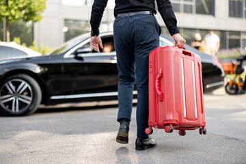Man carries red suitcase to a car, cropped view from below. Concept of chauffeur service and business trips - obrazy, fototapety, plakaty