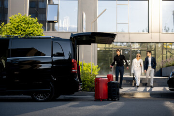 Driver or concierge helps a business couple carry their suitcases to minivan taxi from hotel....