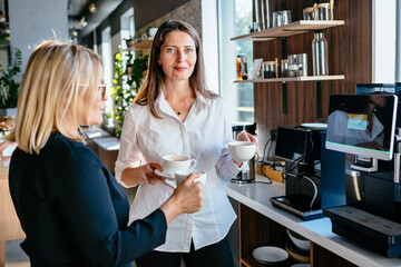 Two mature businesswomen discussing during coffee break. Mature businesswoman on coffee break in office kitchen, holding coffee.