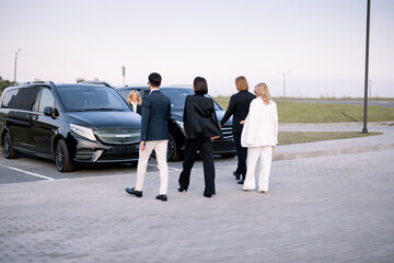 Group of an elegant business people walk together to minivan taxies on a parking lot. Concept of...