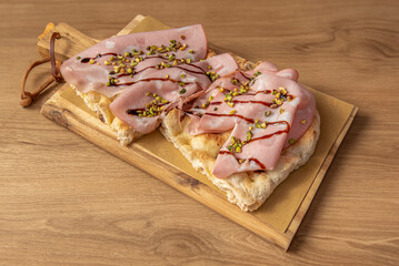 A piece of a traditional pizza of the Roman tradition, known as Pinsa Romana, with Mortadella of Bologna, Gorgonzola and Pistacchio grains, vision from above on wooden table and cutting board 