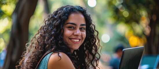 Smiling curly haired girl of Brazilian or Hispanic origin, working or studying outdoors, using a laptop and friendly gaze at the camera.